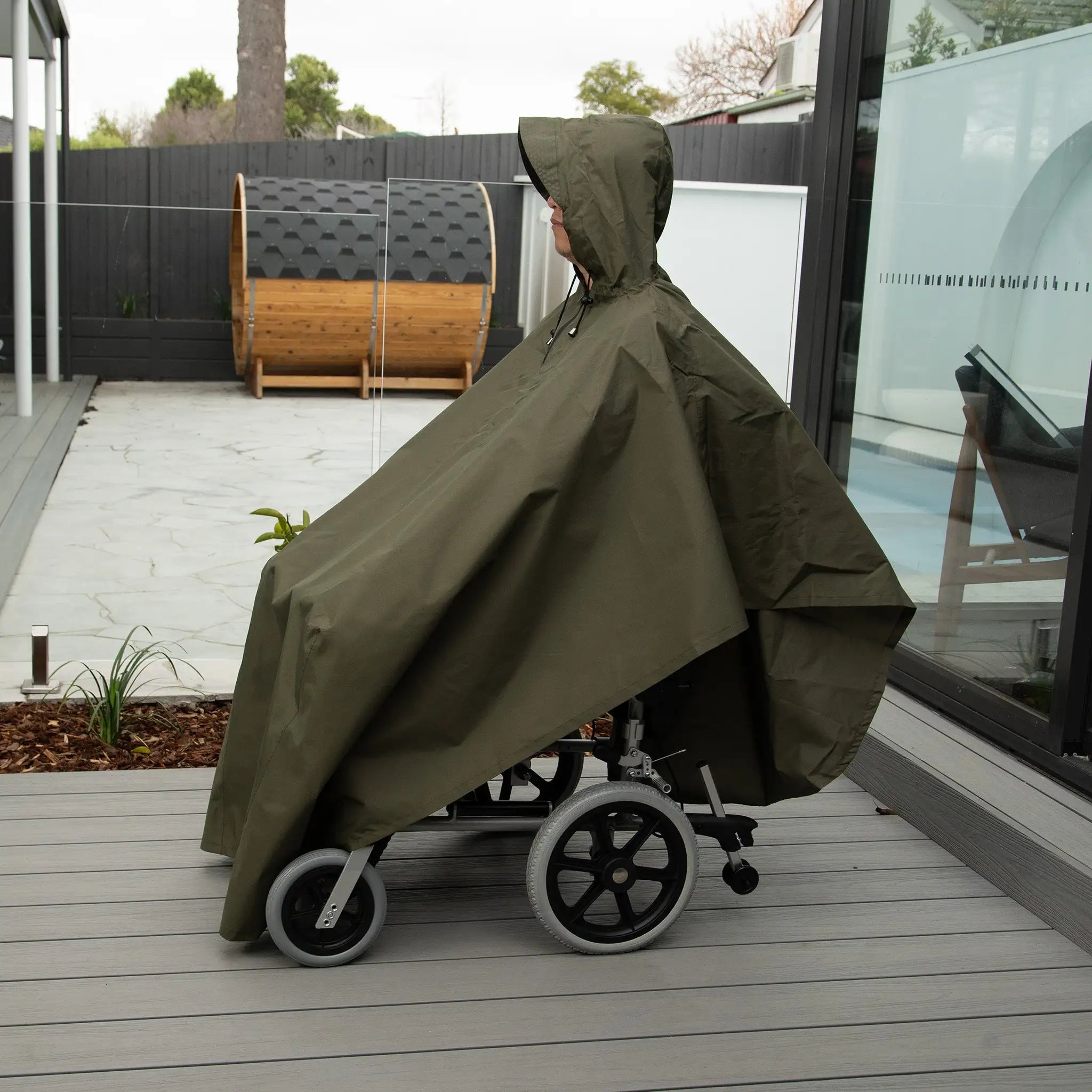 Side view of a person in a wheelchair wearing a waterproof wheelchair poncho in olive green. Designed for full rain and wind protection, this poncho features an adjustable hood and an oversized fit to cover both the user and wheelchair. Made from lightweight, water-resistant fabric, it ensures comfort and mobility in wet weather. Ideal for outdoor activities, travel, and everyday use.