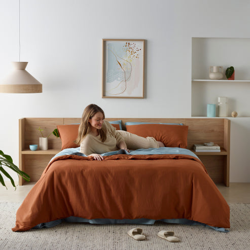 Modern bedroom featuring a burnt orange linen-look water-resistant quilt cover set with matching pillowcases. A woman lounges on the bed, enjoying a book in a cosy, stylish setting with minimalist wooden decor and warm lighting. The waterproof bedding offers comfort and protection while maintaining a chic aesthetic.