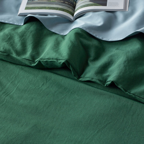 Close-up of a linen-look water-resistant quilt cover set in olive green, layered with a light blue sheet. The soft, textured fabric is displayed with an open book resting on the bedding, highlighting the stylish and functional waterproof material.