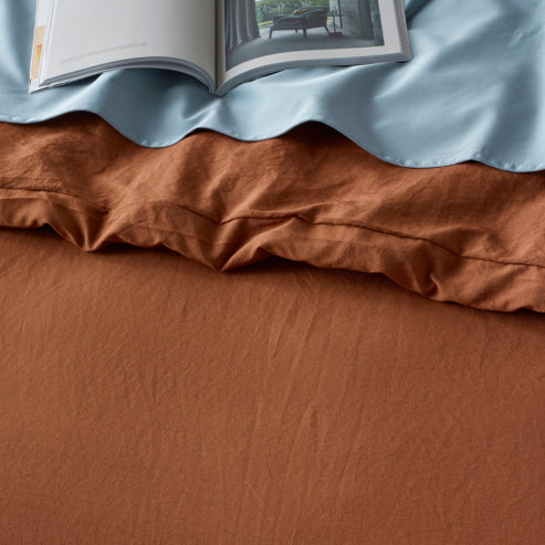 Close-up of a burnt orange linen-look quilt cover set with a soft, water-resistant fabric. The bedding is layered with a light blue sheet and an open book, showcasing its breathable, durable, and stylish design for a cosy and modern bedroom aesthetic.