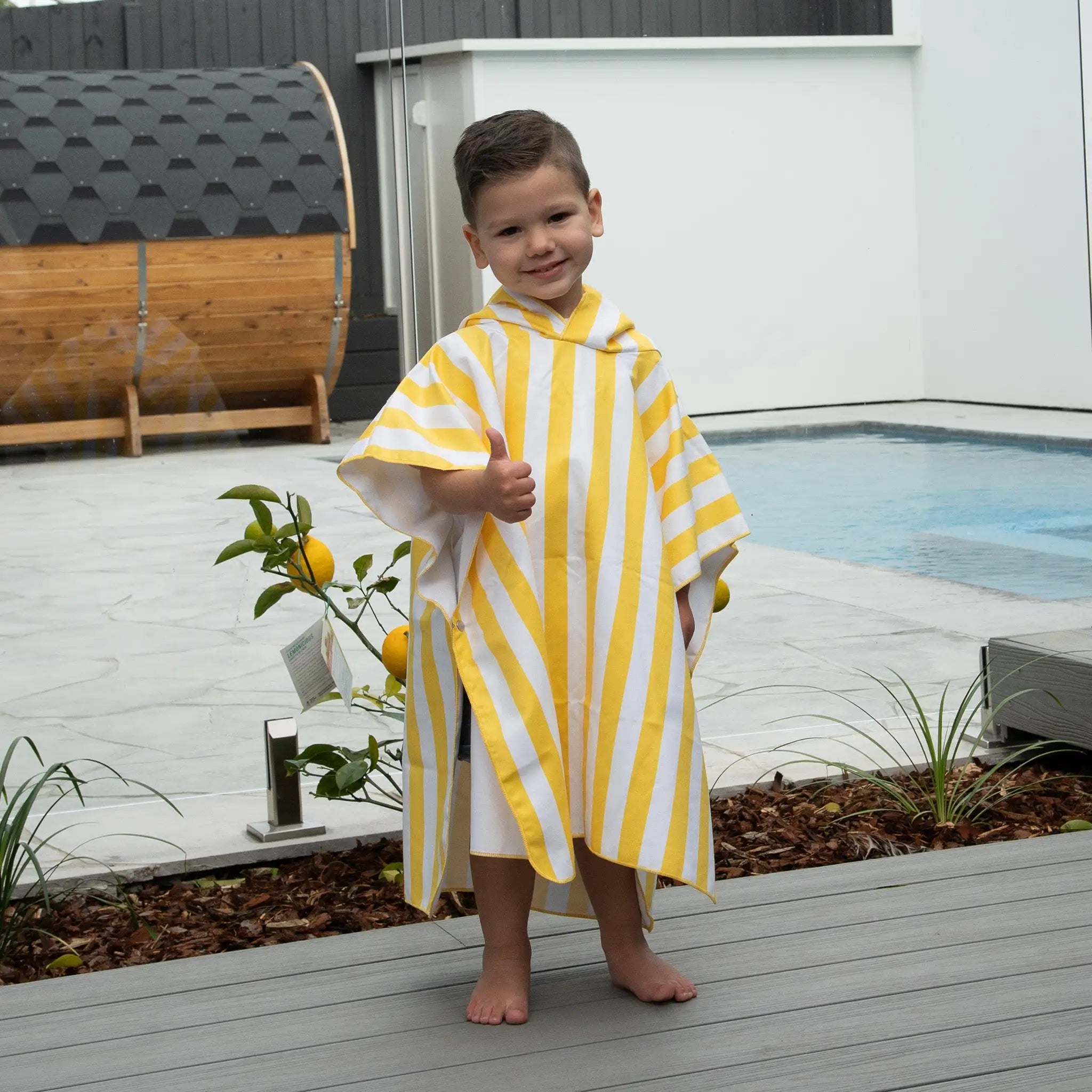 child wearing a StayDry microfibre poncho towel in yellow and white stripes, standing near a swimming pool. Ultra-absorbent, lightweight, and quick-drying towel for beach and bath time.